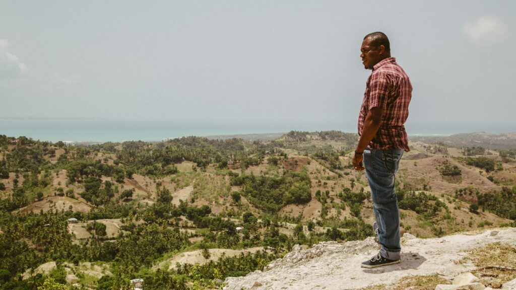 A man stands on a cliff in Haiti, overlooking a scenic landscape with the ocean in the background.